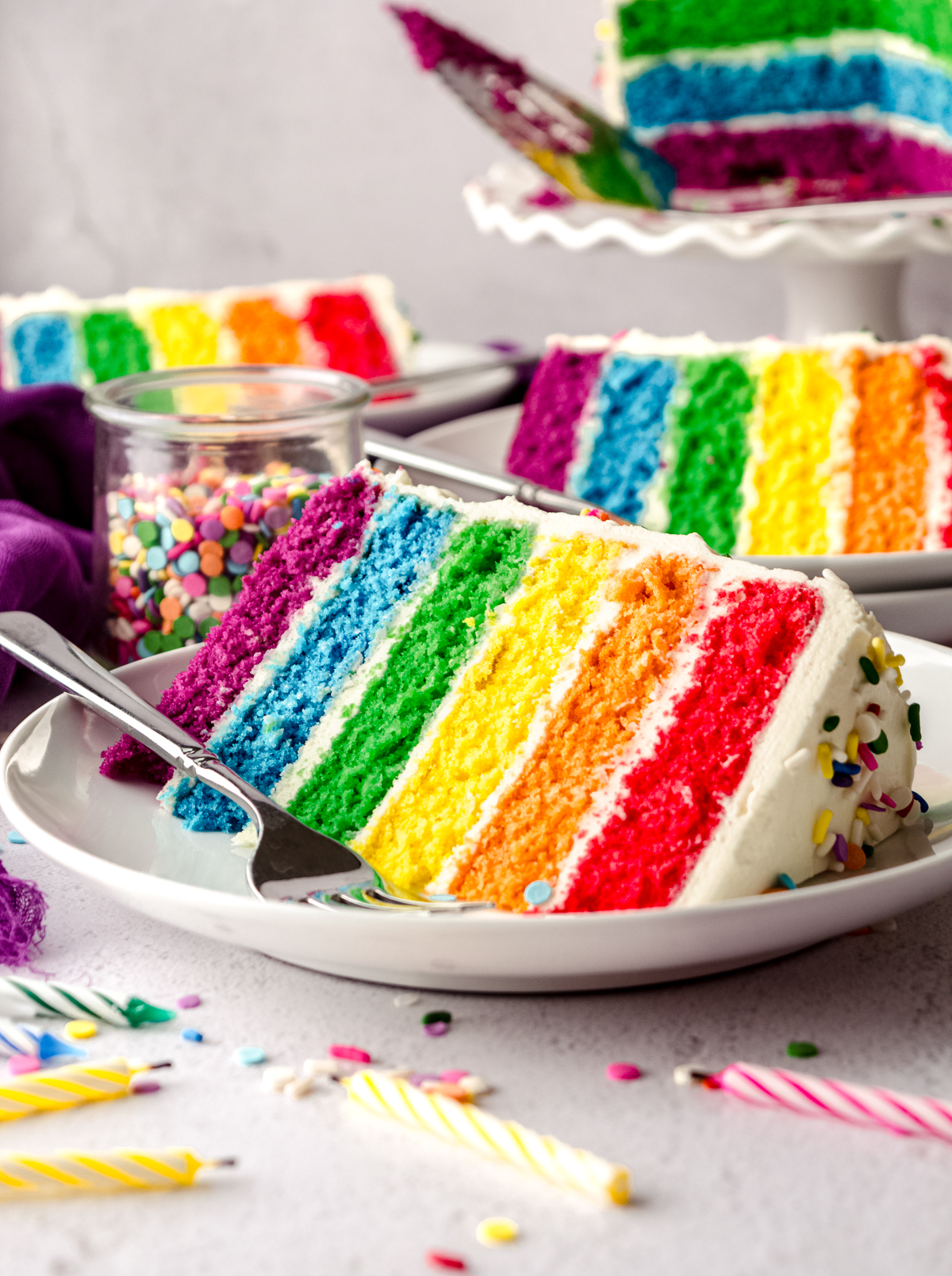 A slice of rainbow layer cake on a plate with a fork and sprinkles around it.