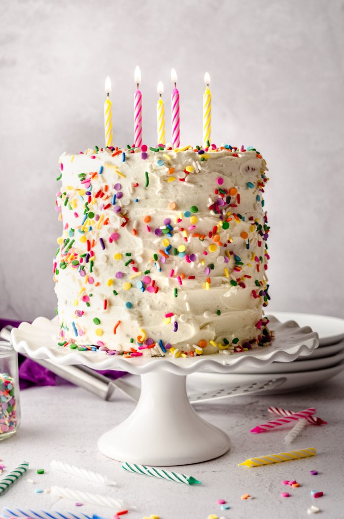 A rainbow layer cake on a cake stand with lit candles in the top of the cake.