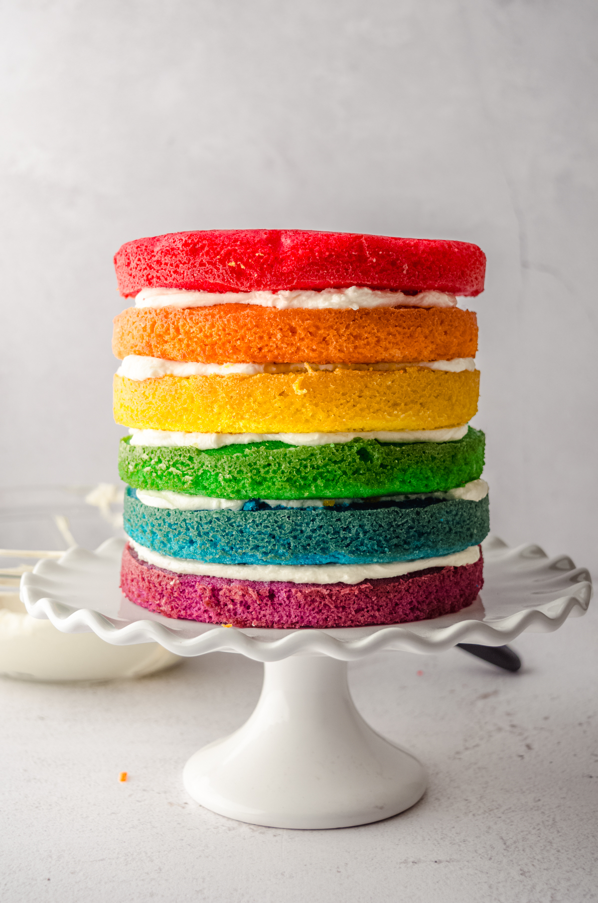 Layers of a rainbow cake stacked with frosting between the layers on a cake stand.