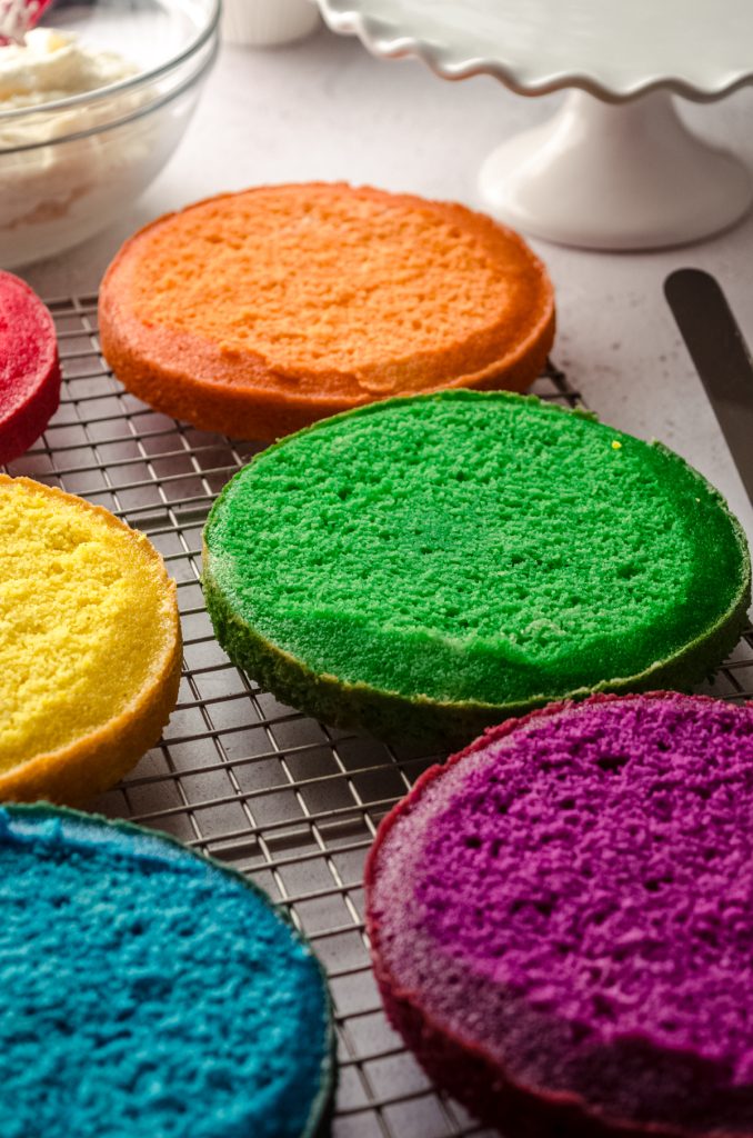 Layers for a rainbow layer cake on a cooling rack that have been trimmed.