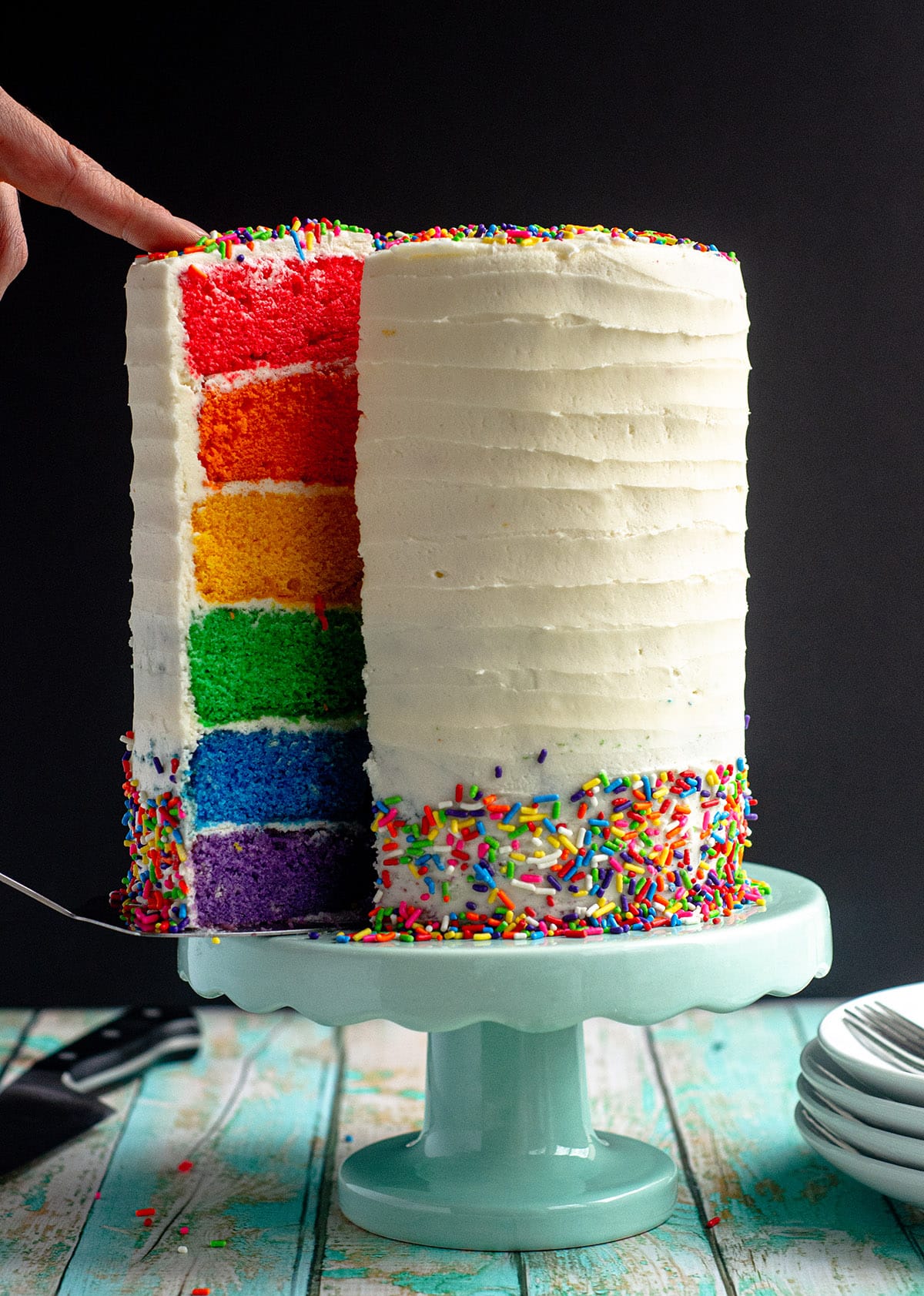 a rainbow cake on a cake stand with a slice being taken out of it