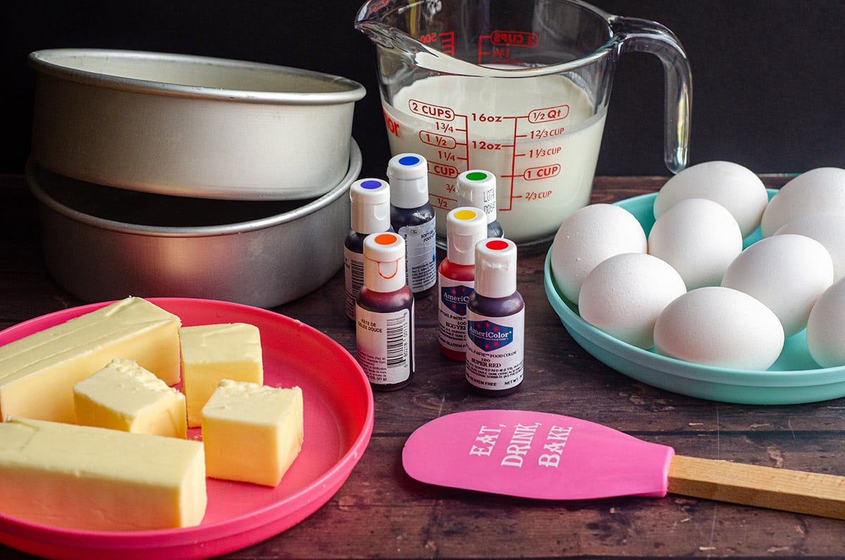 mise en place for a rainbow cake: a plate of butter, cake pans, gel coloring, a spatula, a measuring cup of milk, and a plate full of eggs