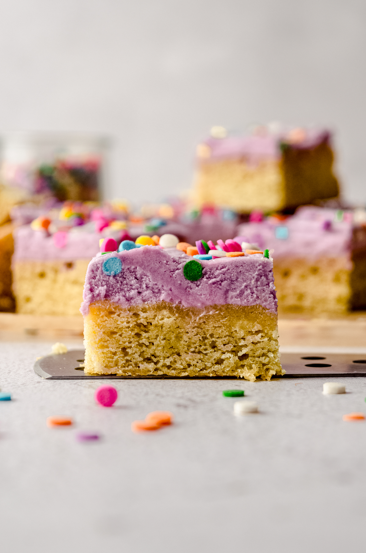 A frosted sugar cookie bar sitting on a serving spatula and you can see other bars in the background.