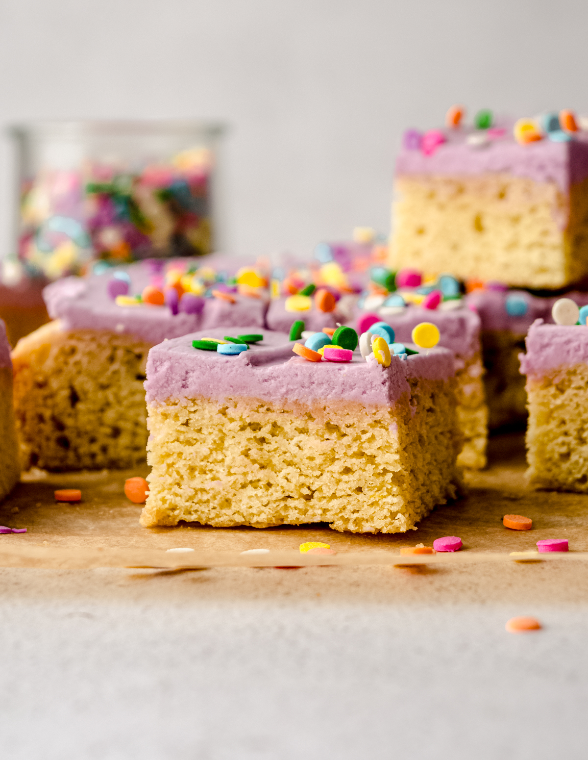 A frosted sugar cookie bar sitting on a piece of parchment paper and you can see other bars in the background.