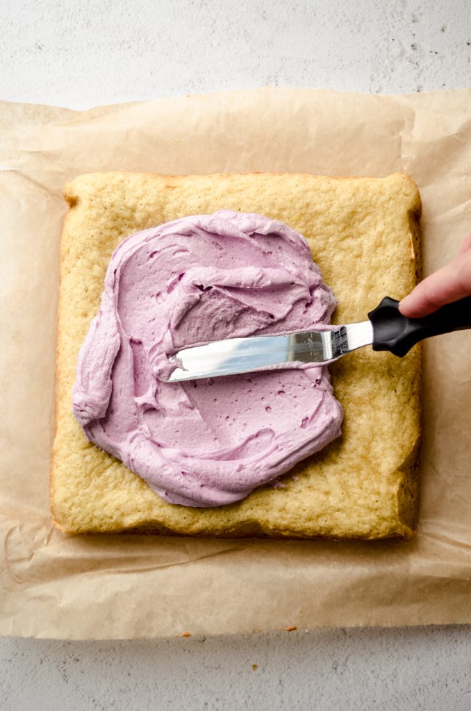 Aerial photo of someone spreading light purple frosting onto a square of sugar cookie bars on a piece of parchment.