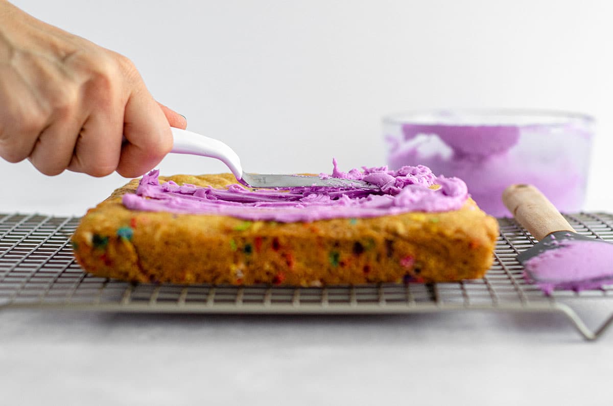 hand spreading purple frosting onto sugar cookie bars with a spatula