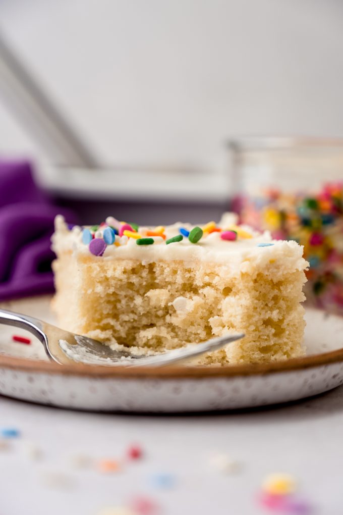 A slice of white sheet cake on a plate with a fork and a bite taken out of the cake.