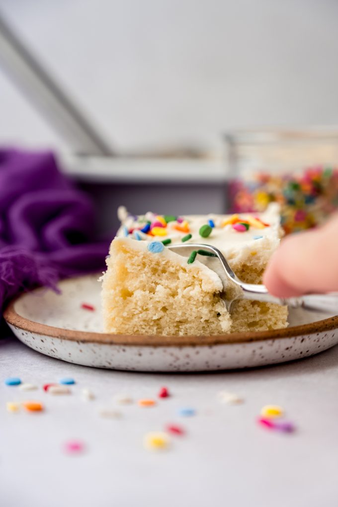 Someone is pushing a fork down into a slice of white sheet cake that's sitting on a plate.