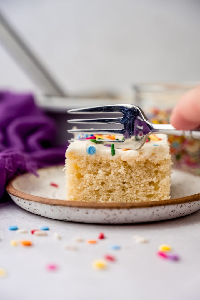 Someone is pushing a fork down into a slice of white sheet cake that's sitting on a plate.