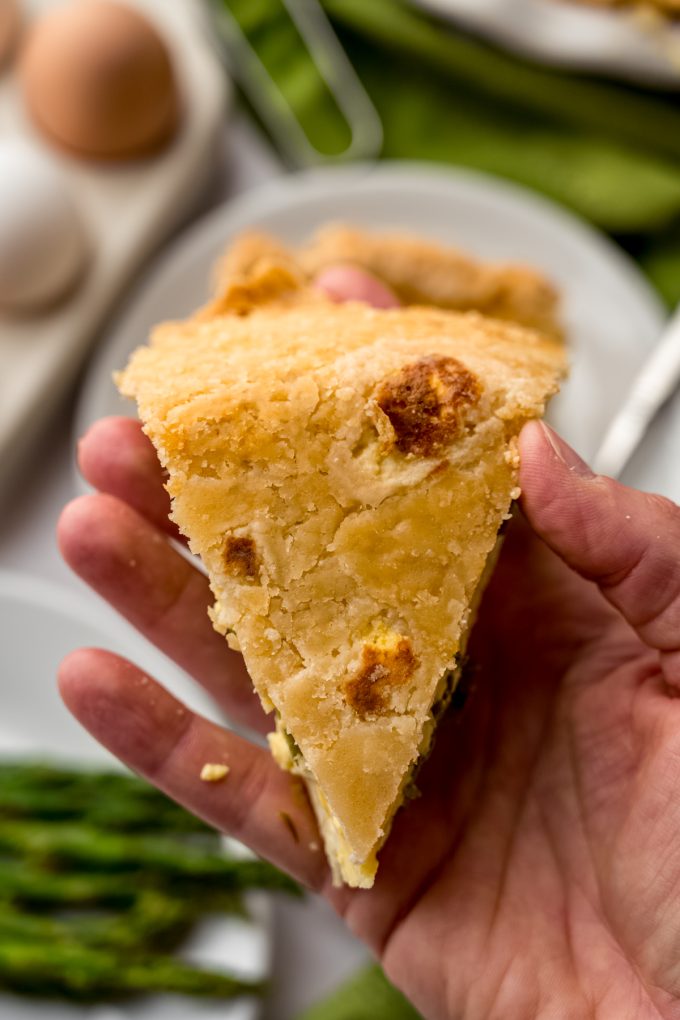 Someone is holding a slice of asparagus quiche upside down to show the flaky crust on the underside.
