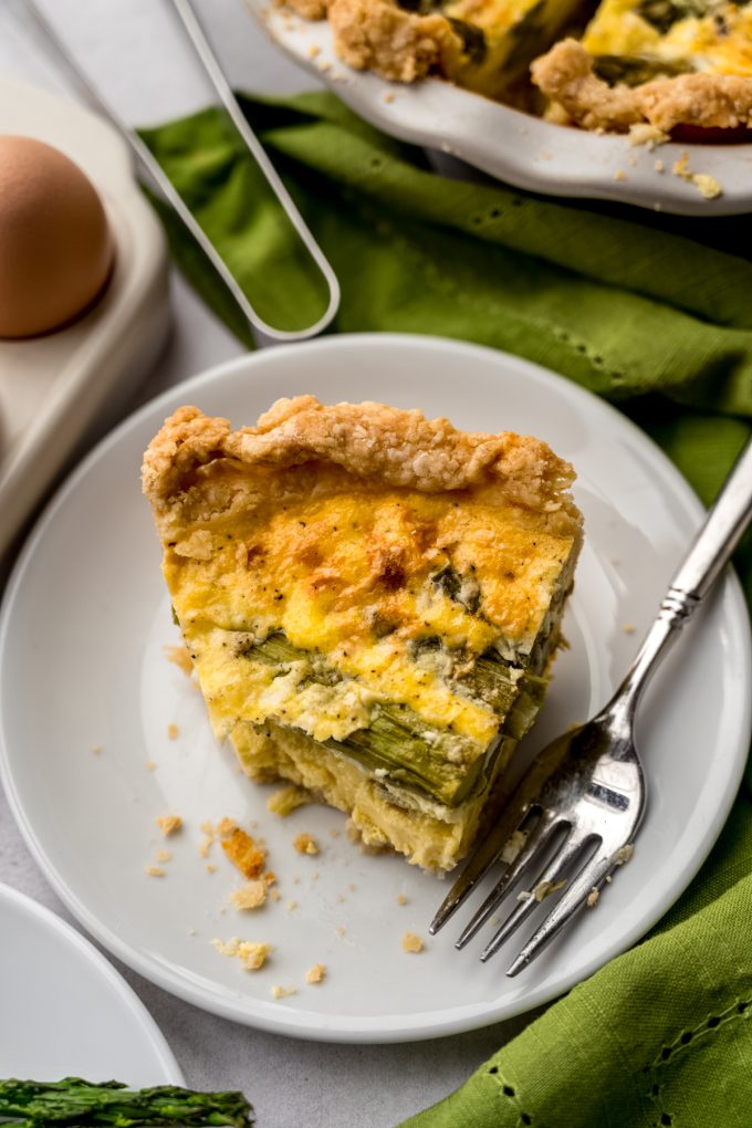 Aerial photo of a slice of asparagus quiche on a plate with a fork with a bite taken out of it.