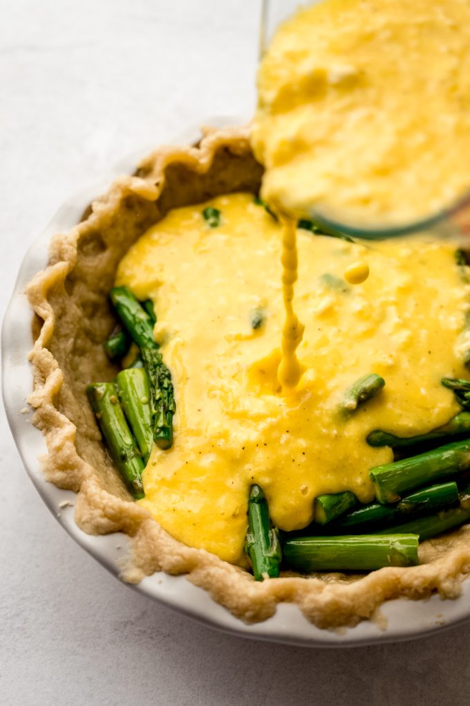 Someone is pouring the egg mixture into a pie plate fitted with a pie crust and asparagus to make quiche.