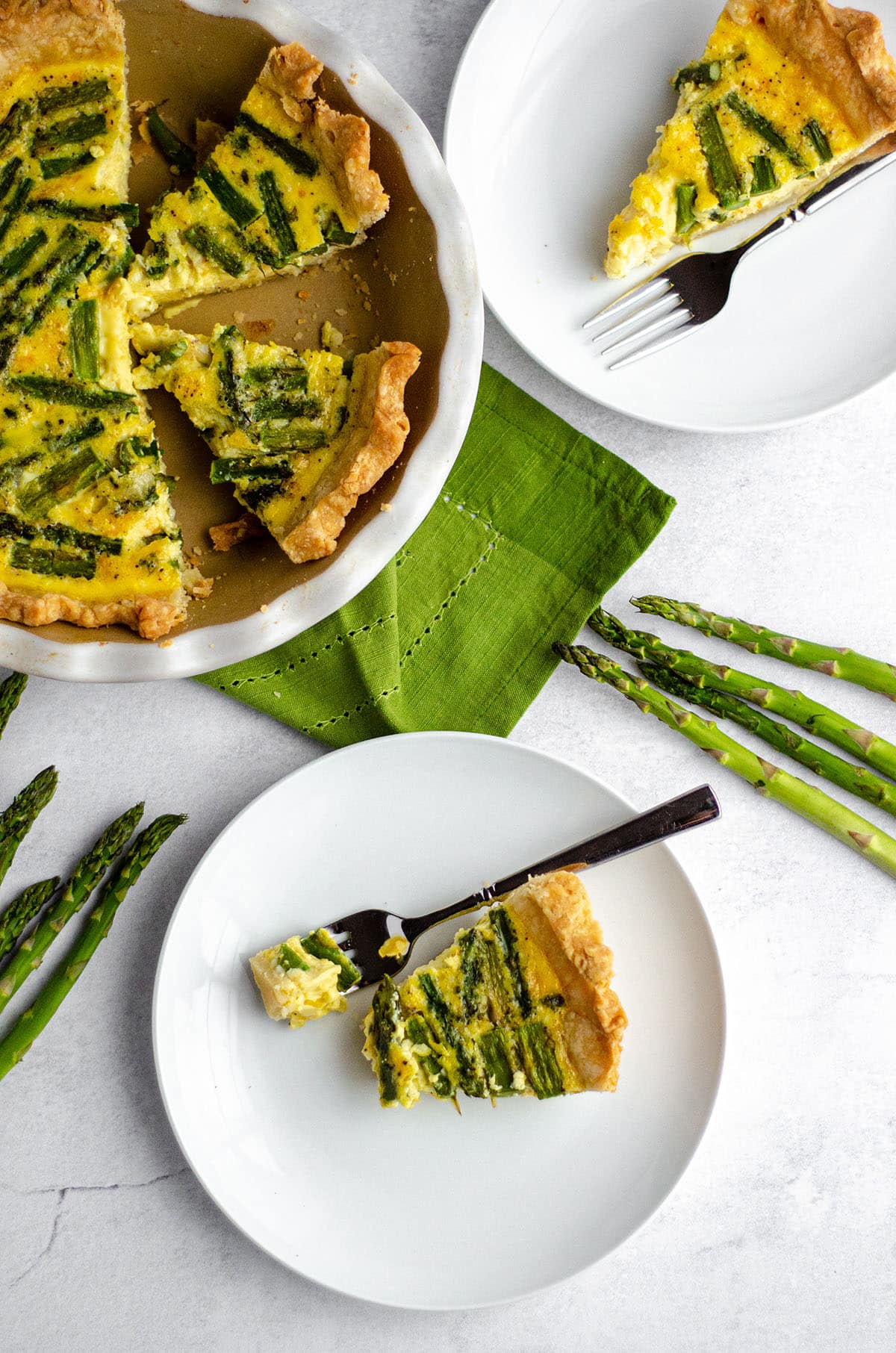 aerial photo of a slice of asparagus quiche on a white plate with a fork and a bite taken out of the slice