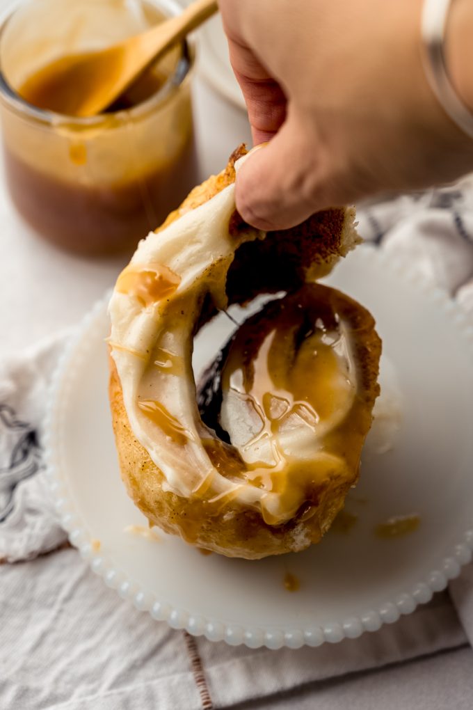 Someone is unraveling a caramel cinnamon roll that's on a plate with a jar of caramel sauce in the background.