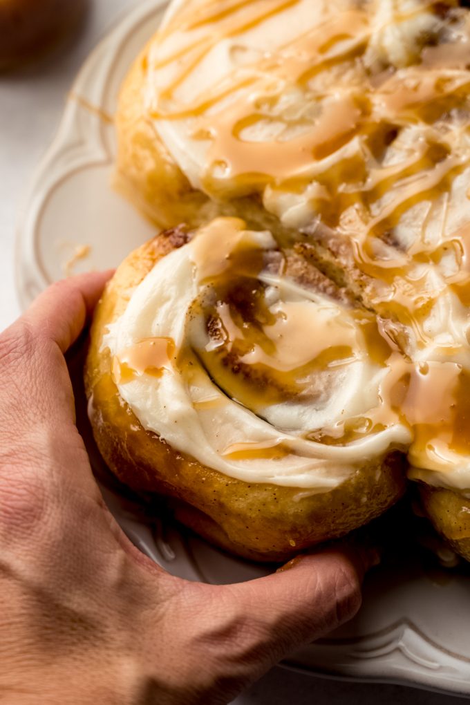 A photo of someone grabbing a roll off of a plate of caramel cinnamon rolls.