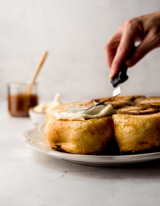 Someone is spreading salted caramel cream cheese frosting onto a plate of caramel cinnamon rolls.