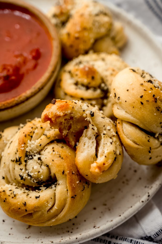 A plate of homemade garlic knots and one has a bite taken out of it.