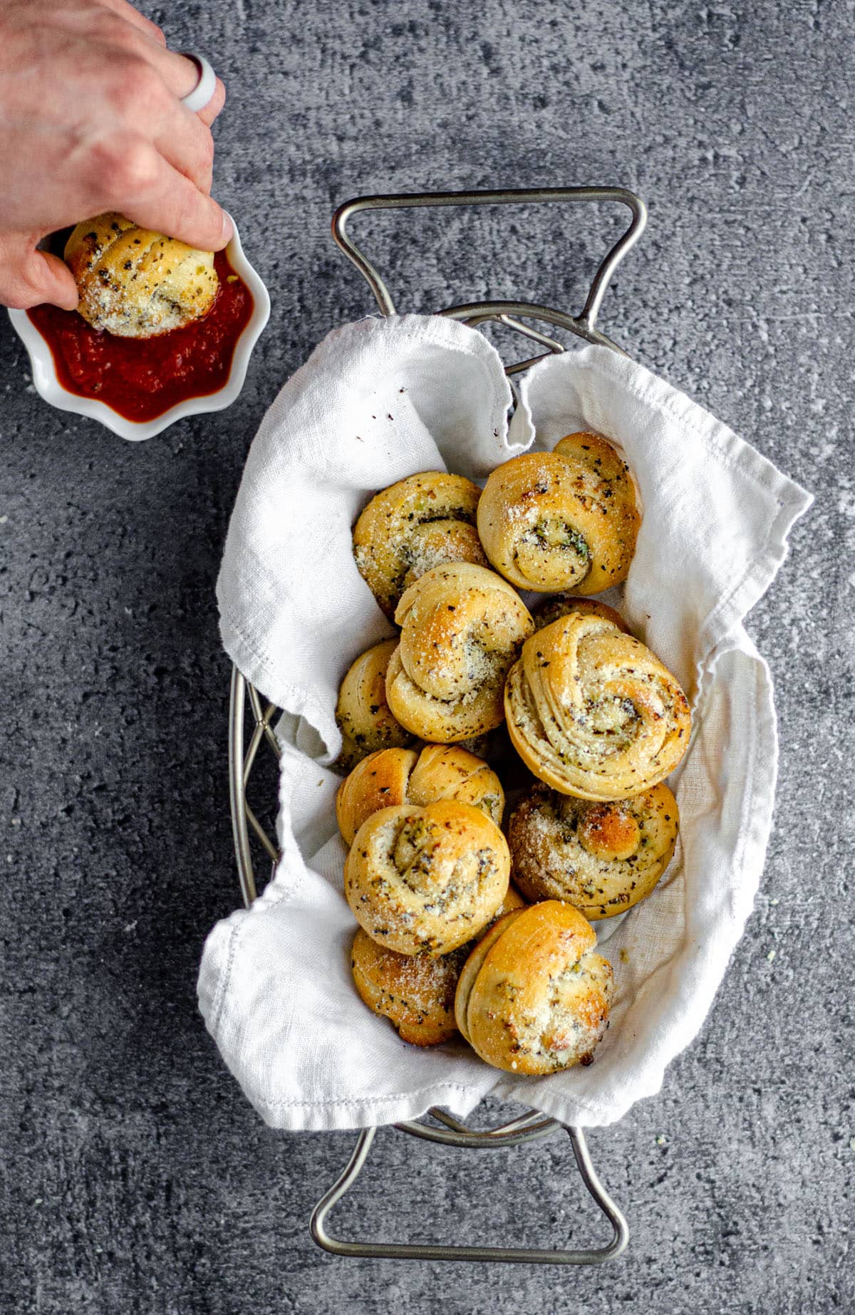 aerial photo of homemade garlic knots in a basket with someone dipping one into marinara off to the side