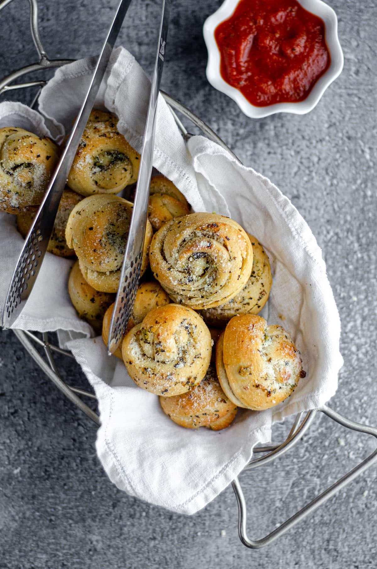 aerial photo of homemade garlic knots in a basket with metal tongs and a cup of marinara