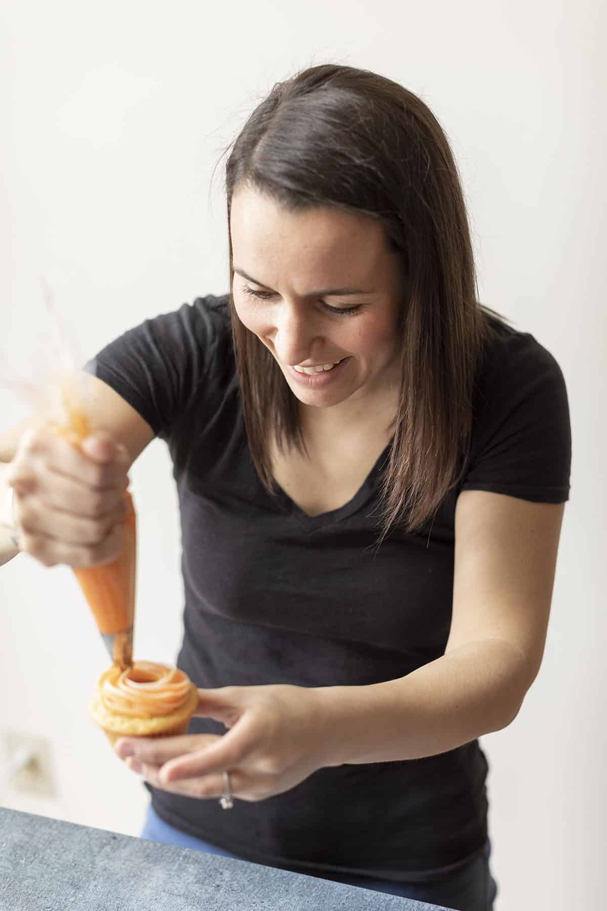 a woman piping orange creamsicle frosting onto an orange creamsicle cupcake