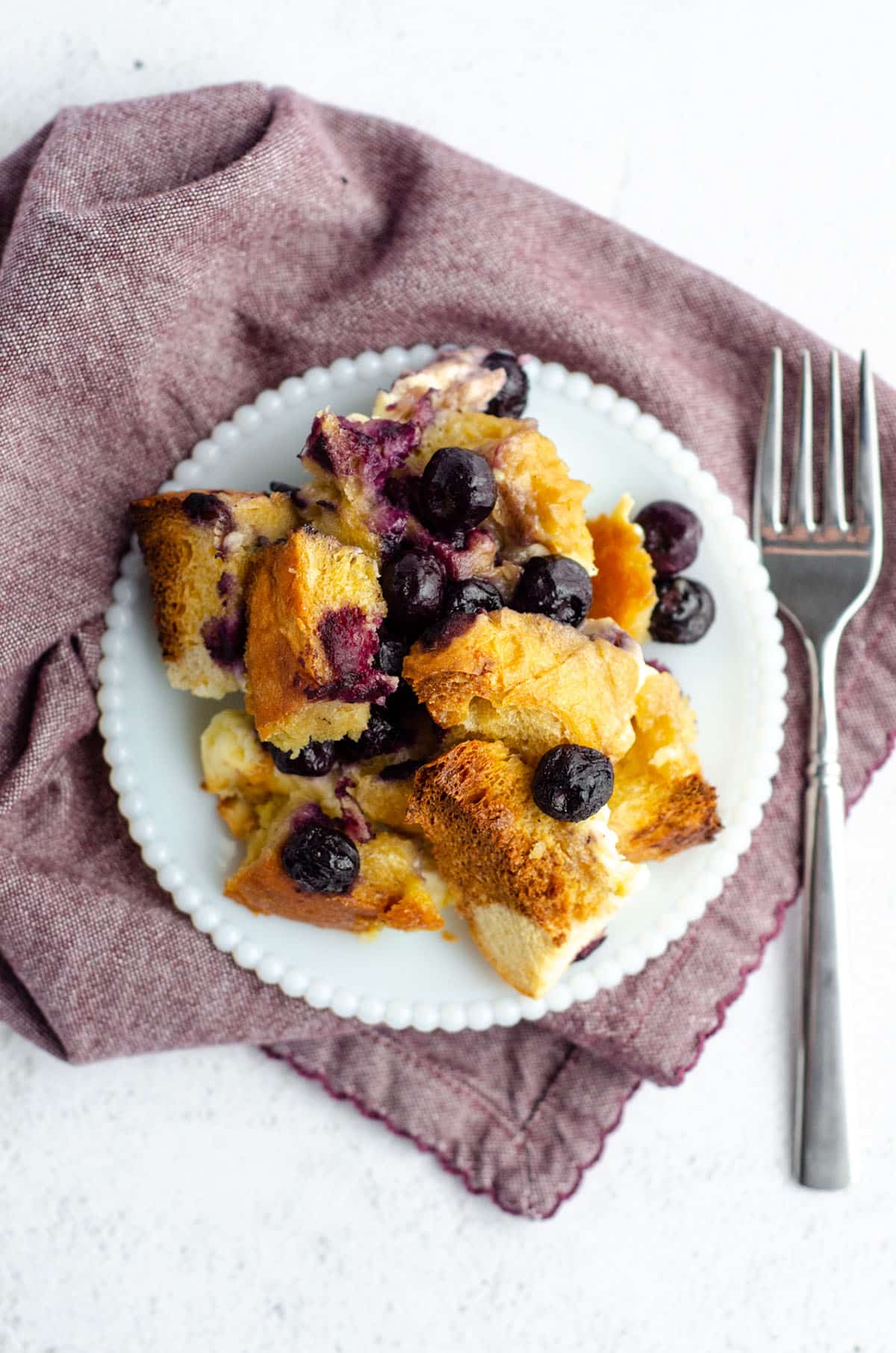 aerial photo of baked blueberry french toast sitting on a white plate with a fork and a plum colored kitchen towel