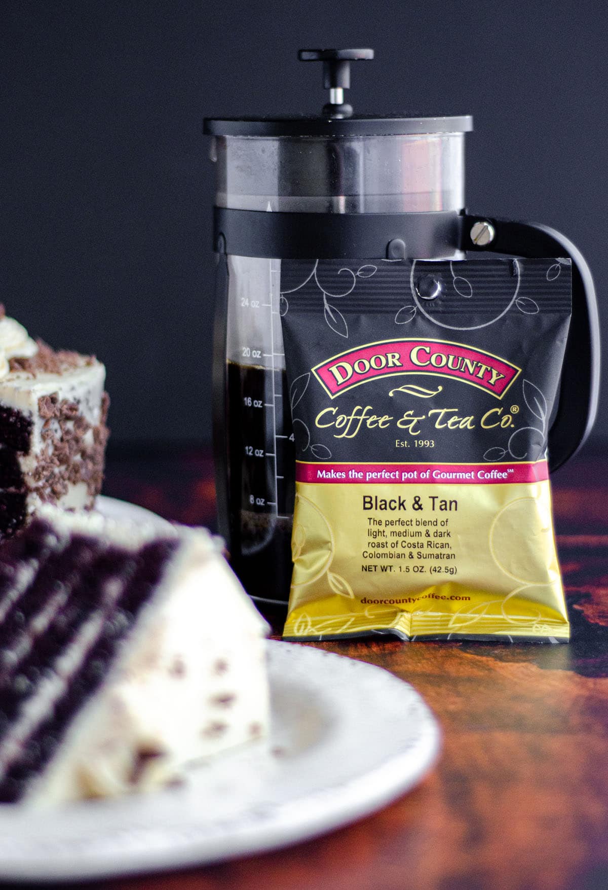 bag of door county coffee leaning up against a french press with a slice of mocha cake in the foreground