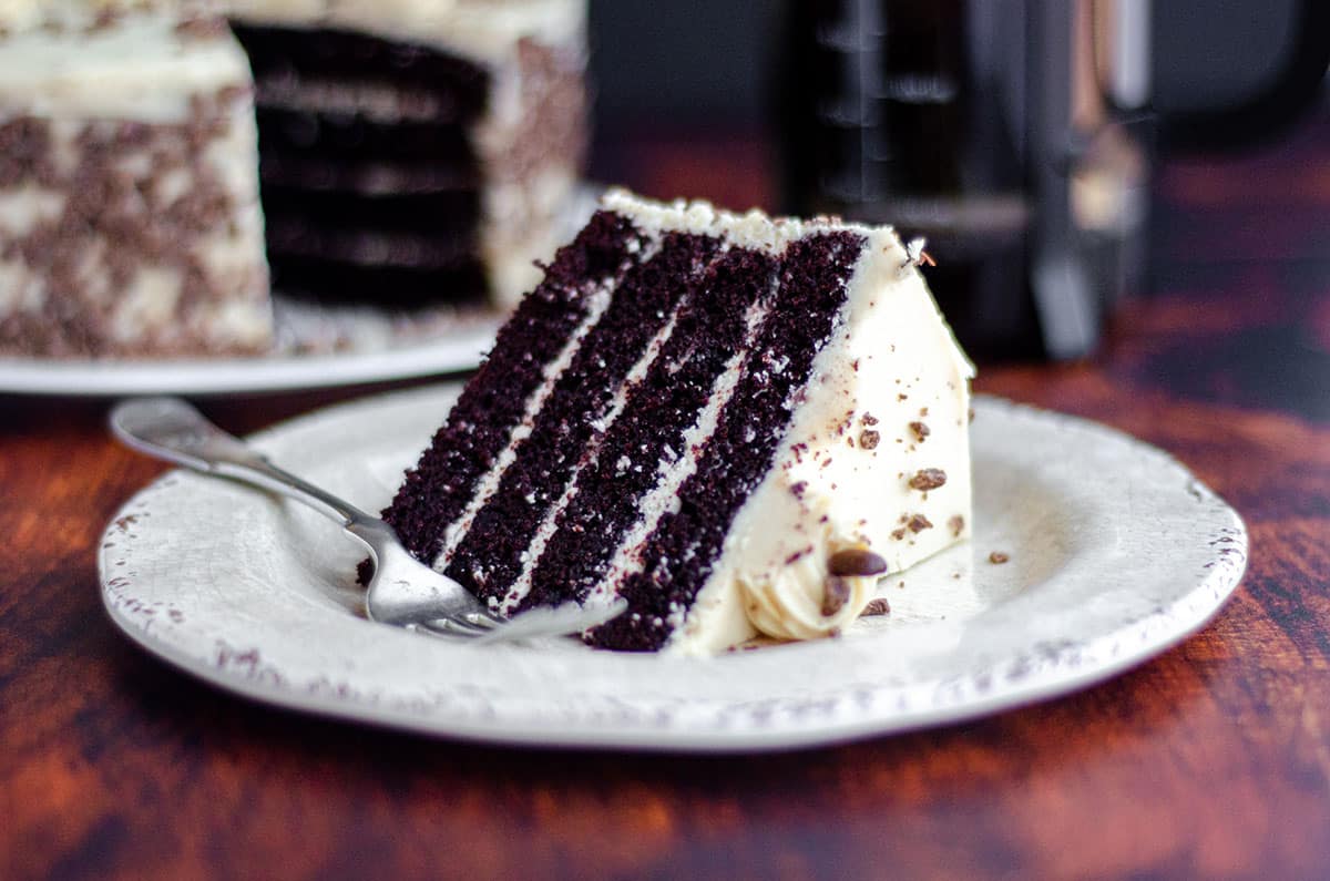 slice of mocha cake with coffee buttercream on a cream colored plate with a fork and a full cake and french press of coffee in the background