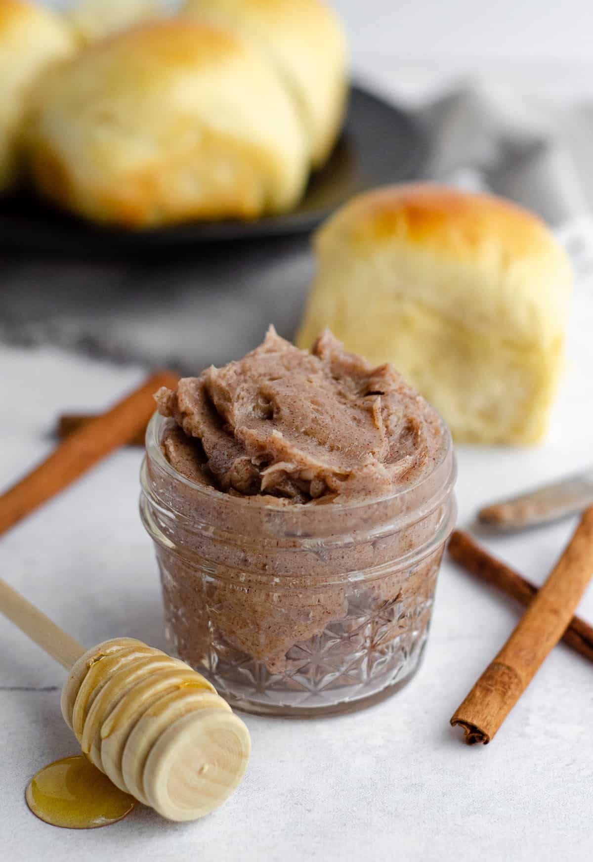 cinnamon honey butter in a jar with cinnamon sticks and a honey dipper  and a plate of rolls in the background