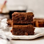 Two coffee brownies stacked on top of each other on a plate with coffee in the background.
