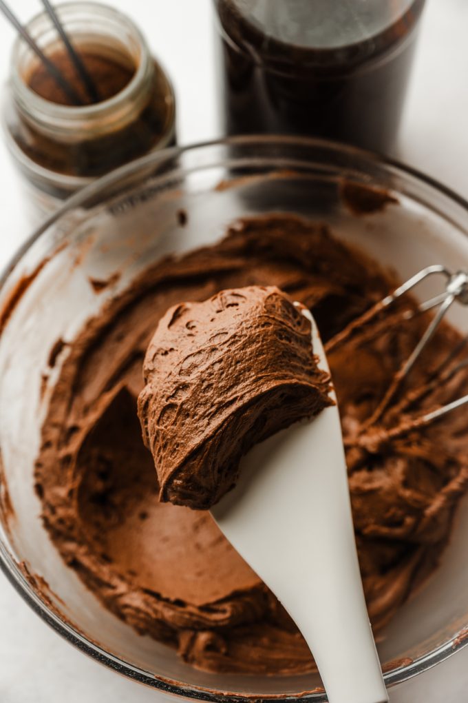 Someone is holding a spatula with mocha frosting on it over a bowl of more frosting.