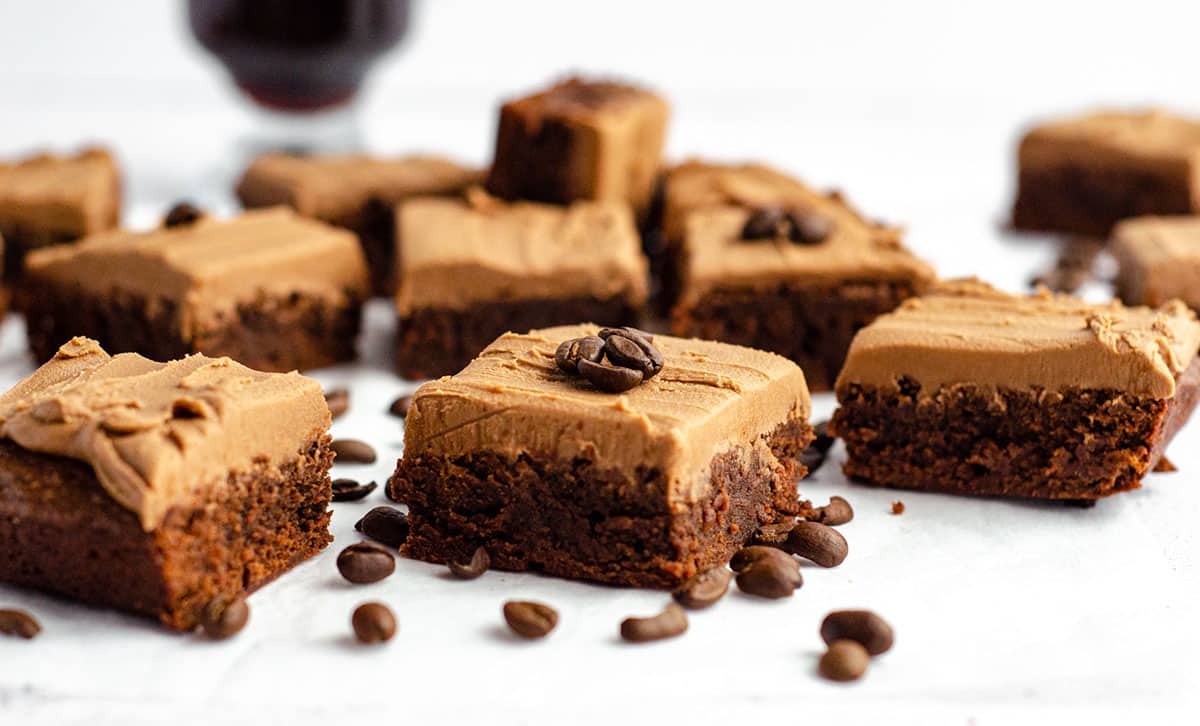 coffee brownies with mocha frosting sliced and on a white surface with coffee beans scattered around it