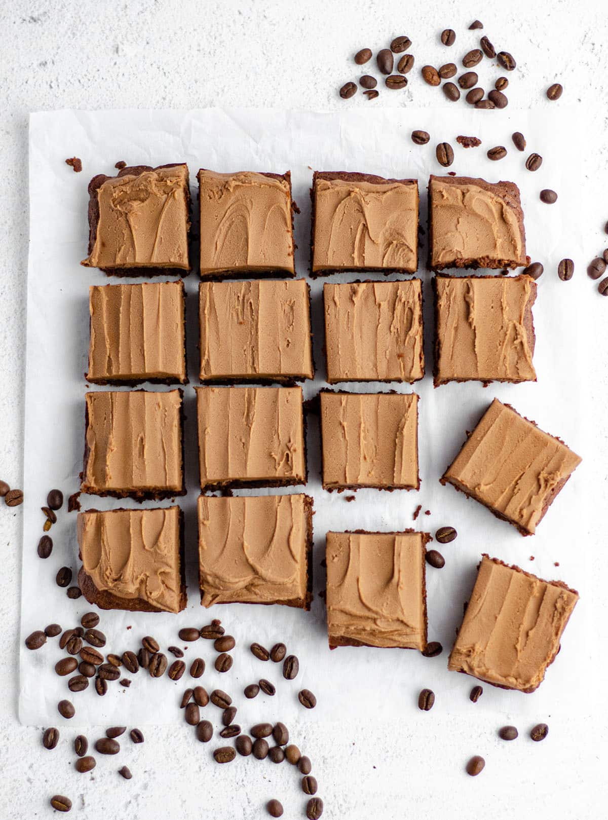 aerial photo of coffee brownies with mocha frosting sliced and sitting on white parchment with coffee beans scattered around