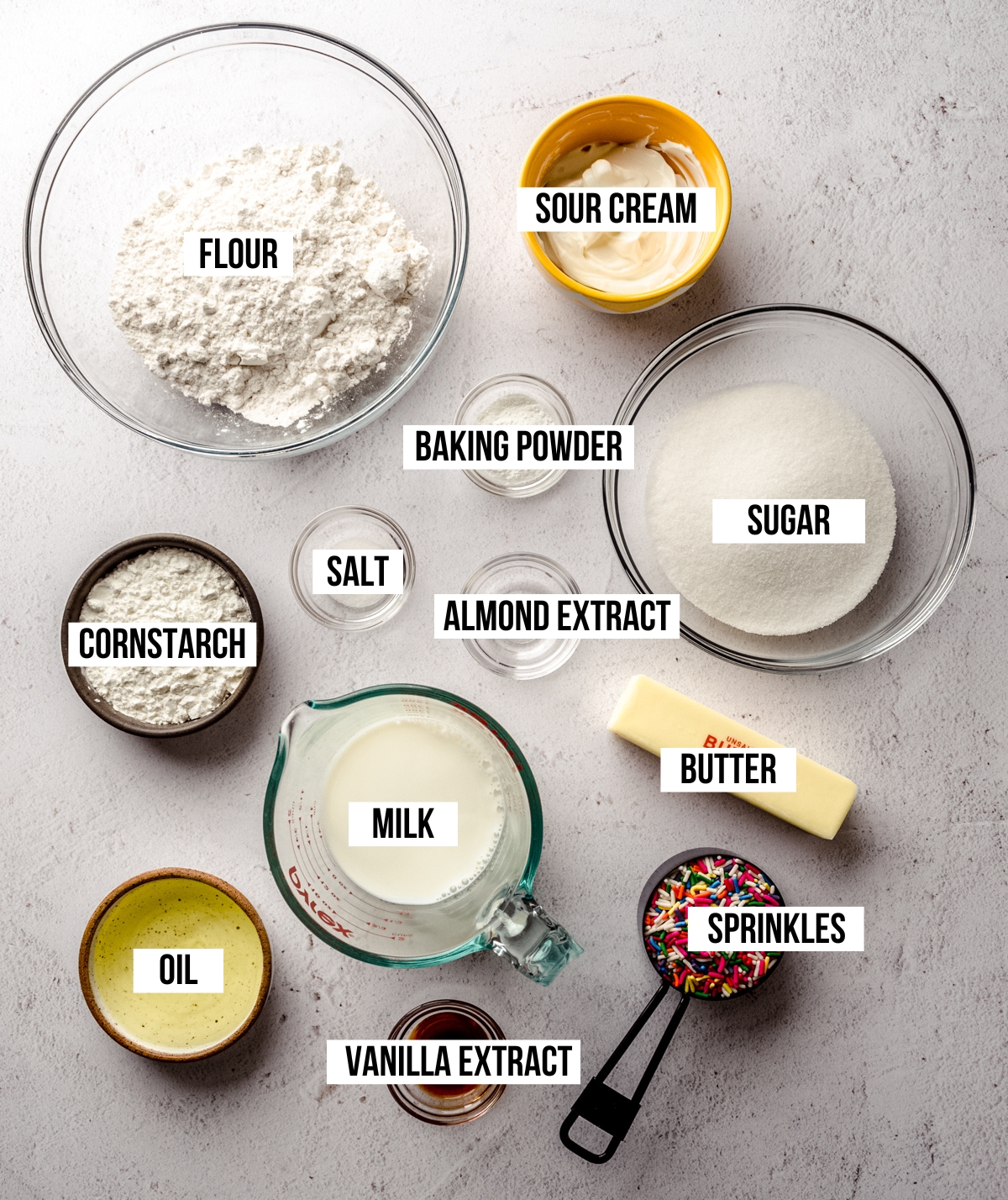 Aerial photo of ingredients for funfetti cupcakes on a surface with text overlay labeling each ingredient.