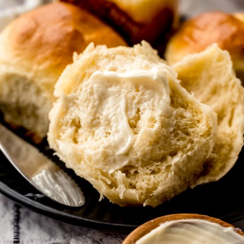 A yeast roll that has been split in half and buttered sitting on a plate with butter in the foreground and more rolls in the background.