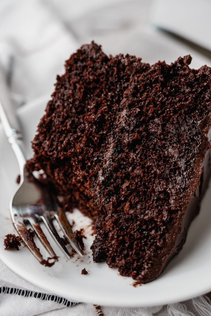 A slice of chocolate layer cake on a plate with a bite taken out of it with a fork.