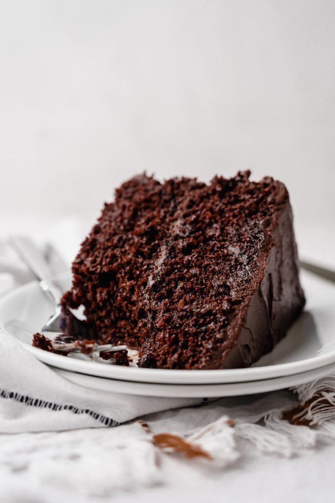 A slice of chocolate layer cake on a plate with a bite taken out of it with a fork.