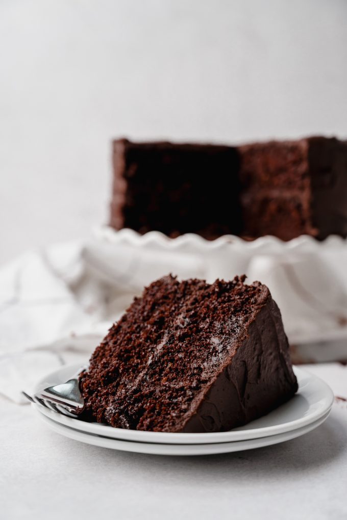 A slice of chocolate cake on a plate with a cake on a cake stand in the background.