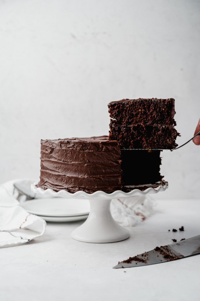 Someone is using a cake server to remove a slice of chocolate cake from a cake on a cake stand.