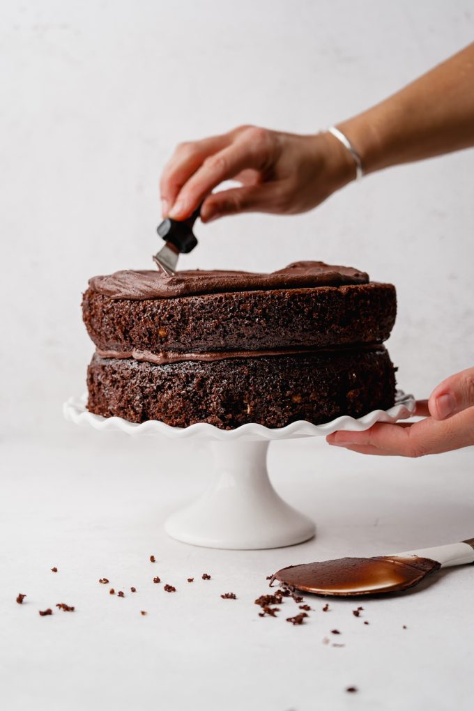 Someone is spreading chocolate frosting onto a chocolate layer cake that is on a fluted cake stand.