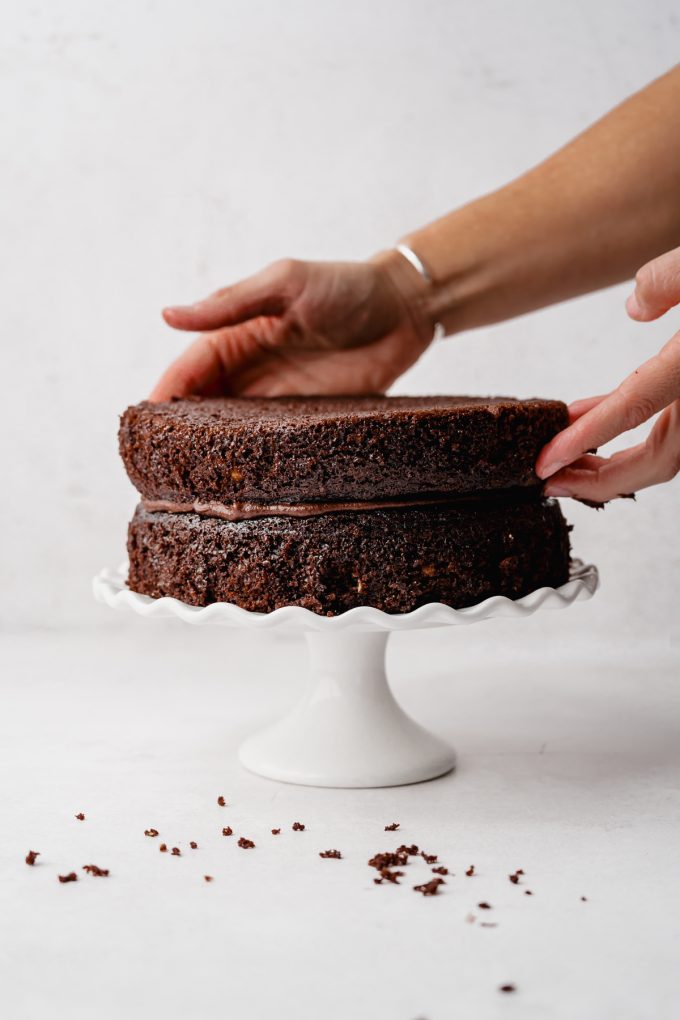Someone is placing a top layer onto a chocolate cake layer that is on a fluted cake stand.