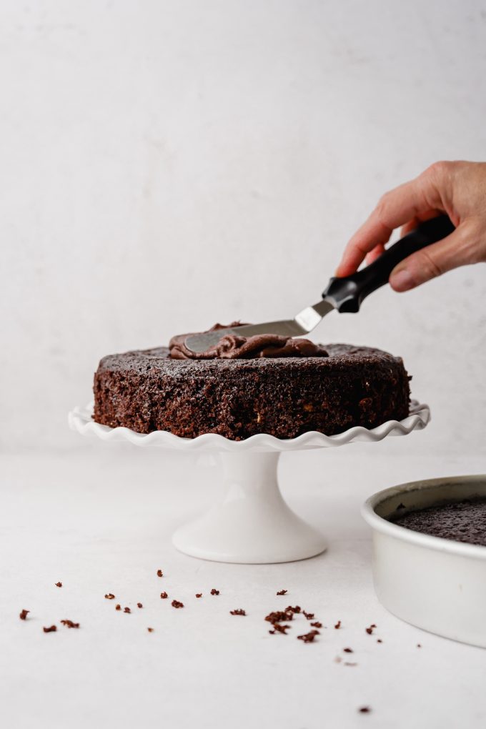 Someone is spreading chocolate frosting onto a cake layer that is on a fluted cake stand.