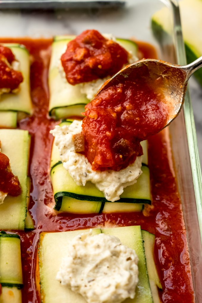 Someone is spooning sauce onto unbaked zucchini ravioli in a baking dish.