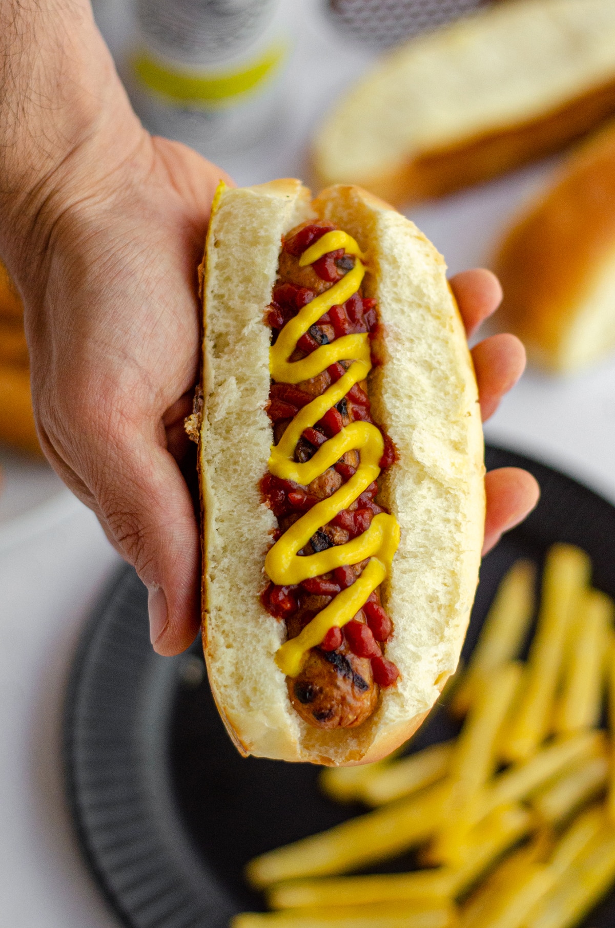 hand holding a hot dog in a homemade hot dog bun with a squirts of ketchup and mustard