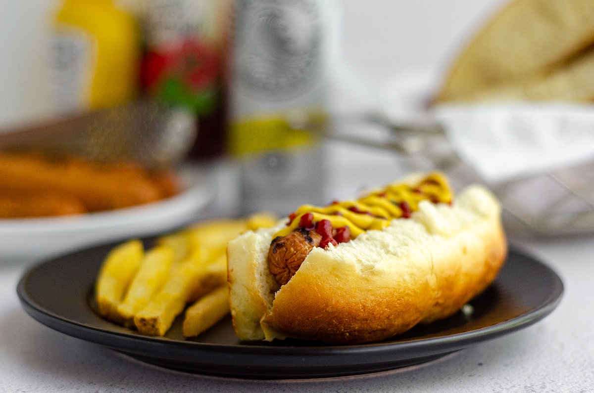 hot dog in a homemade hot dog bun with a squirts of ketchup and mustard sitting on a black plate with french fries