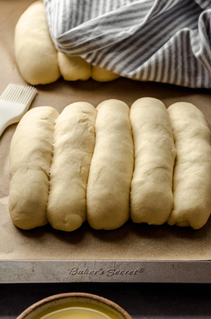 Homemade hot dog buns on a baking sheet that have risen and are ready to bake.