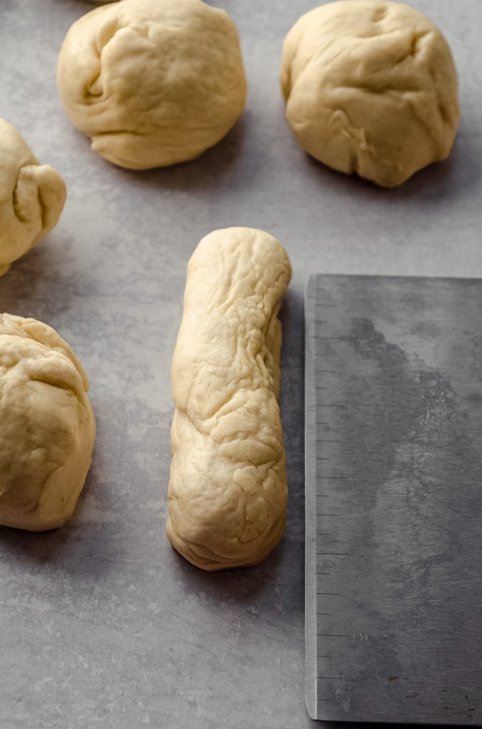 A piece of dough to make a hot dog bun on a surface and it is being measured with a graduated bench scraper.