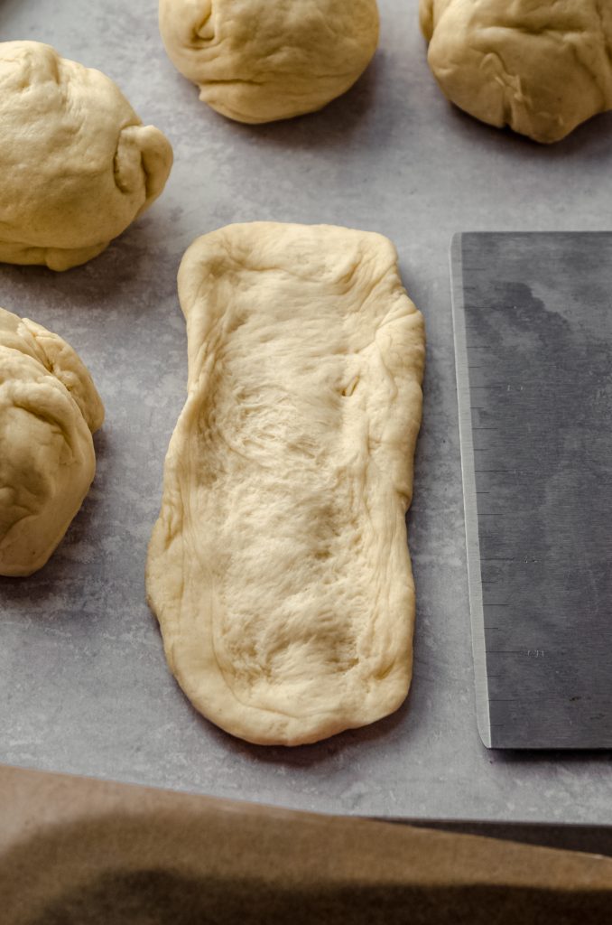 A piece of dough to make a hot dog bun on a surface and it is being measured with a graduated bench scraper.