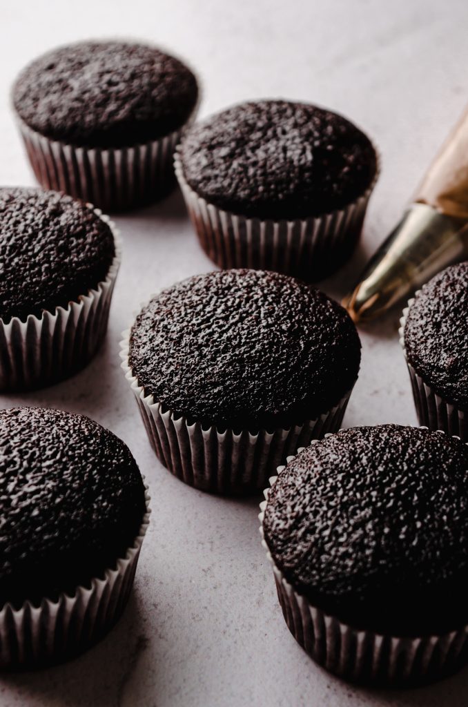 Unfrosted chocolate cupcakes sitting on a surface with a piping back in the background.