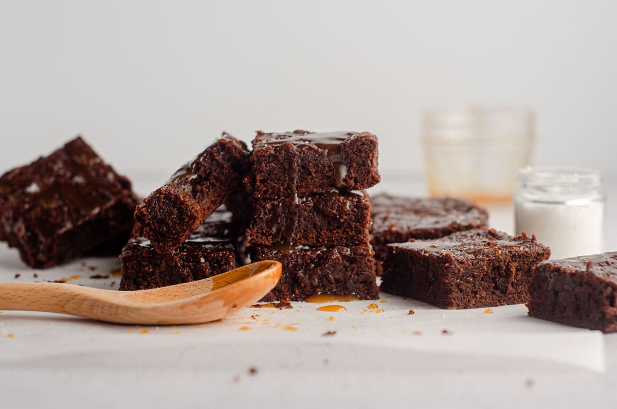 stack of salted caramel brownies with a wooden spoon that has caramel sauce dripping off of it in the foreground