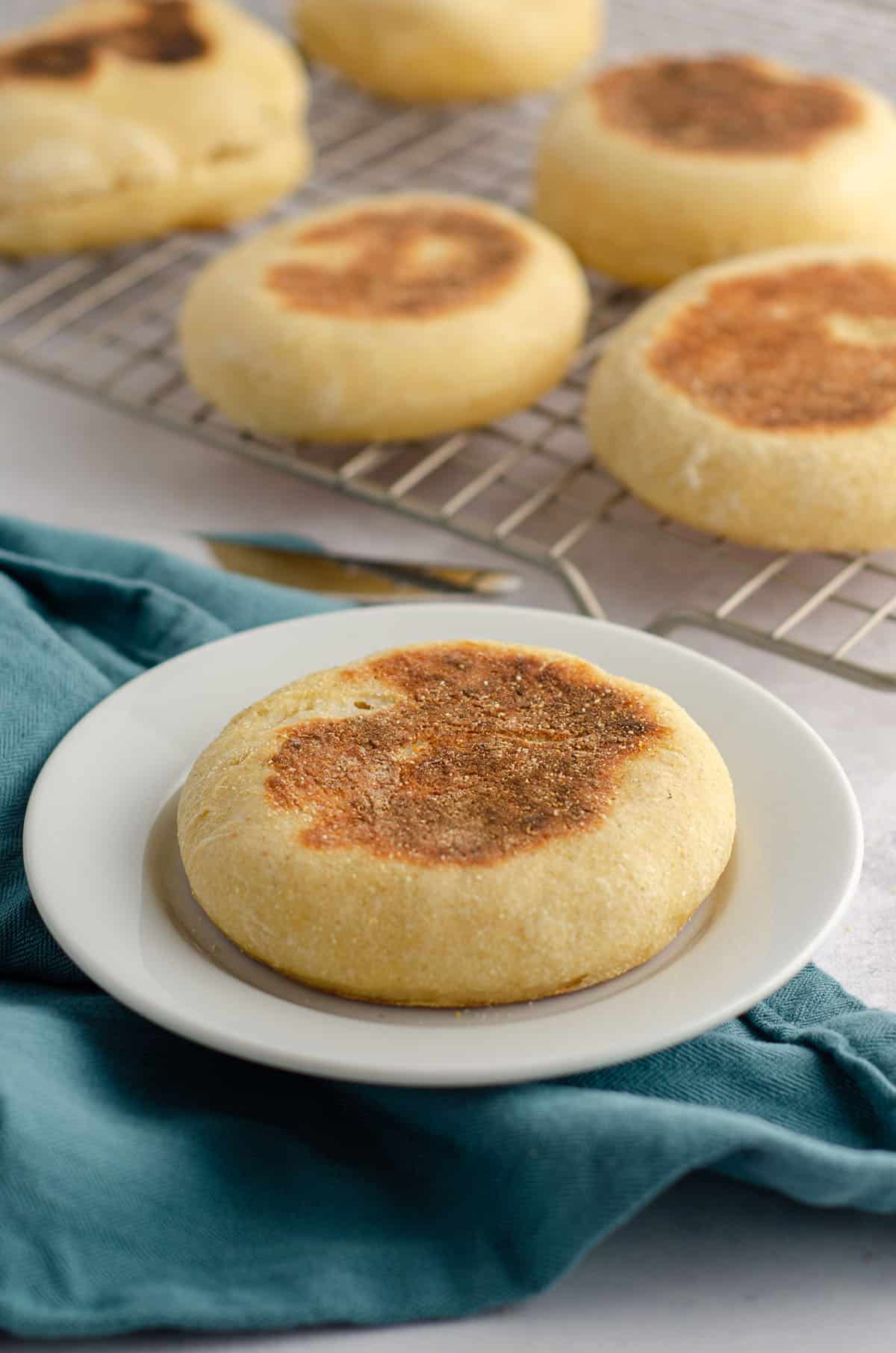 sourdough english muffins on a cooling rack in the background of a sourdough english muffin sitting on a white plate